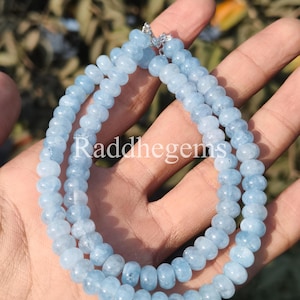 Op de afbeelding: Een lichtblauwe aquamarijn edelsteenketting. De ketting is gemaakt van kleine, afgeronde kralen die aan elkaar zijn geregen. De tekst "Raddhegems" is zichtbaar in de afbeelding. De ketting wordt gepresenteerd tegen een vage achtergrond.