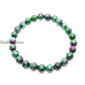 Peut inclure: Un bracelet de perles composé de pierres précieuses rondes vertes et violettes. Les perles sont enfilées ensemble, formant une forme circulaire. Le texte "Raddigems" est visible sur le côté gauche de l'image.