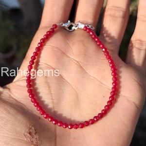 Peut inclure: Un bracelet délicat en pierres précieuses rouges avec un fermoir argenté. Le bracelet est composé de petites perles rondes et facettées. Le bracelet est présenté sur une main. Le texte "Rahegems" est visible.