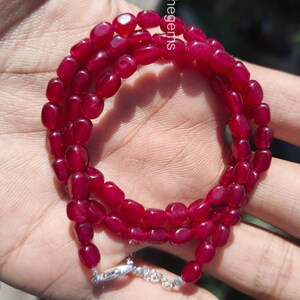 Op de afbeelding: Een ketting bestaande uit drie strengen gepolijste, ovaalvormige, dieprode edelstenen. De ketting is vastgemaakt met een zilveren sluiting. De edelstenen hebben een levendige, rijke kleur en de ketting wordt gepresenteerd tegen een neutrale achtergrond.