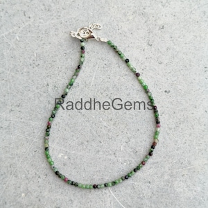 May include: A delicate beaded bracelet featuring small, round green, black, and red gemstones. The bracelet is secured with a silver-colored clasp. The text "RaddheGems" is visible on the bracelet.