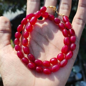 May include: A double-strand necklace featuring oval-shaped, deep red beads. The beads are strung together with a gold-colored chain and clasp. The necklace is displayed on a hand.