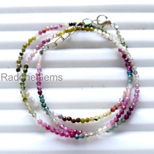 以下が含まれることがあります： 小さなカラフルな宝石ビーズが付いた3つの繊細なネックレス。ビーズはピンク、グリーン、ブルー、ホワイトの色合いです。ネックレスはシルバーの留め金が付いており、重ね着けされています。「Radche Gems」の文字が見えます。