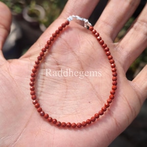 May include: A red beaded bracelet with a silver clasp. The bracelet is made of small, faceted beads. The text "Raddhegems" is visible on the image. The bracelet is displayed on a hand.
