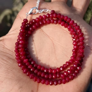 May include: A three-strand bracelet made of small, faceted, deep red beads. The bracelet has a silver-coloured clasp and chain. The beads are arranged in a circular pattern.