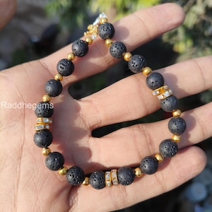May include: A beaded bracelet featuring black lava stone beads, interspersed with gold-colored beads and sections of clear, sparkling crystals. The bracelet is strung on a gold-colored chain and is designed for wrist wear.