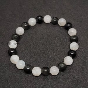 以下が含まれることがあります： 黒と白の丸い宝石が交互に並んだビーズブレスレット。 ブレスレットは研磨された石でできており、濃い灰色の表面に表示されています。 ブランド名「Raddhegins」が左下に表示されています。