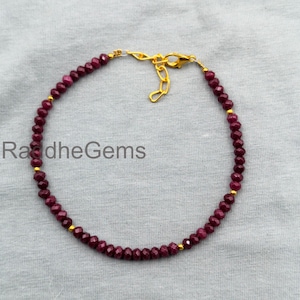 May include: A delicate bracelet featuring small, round, deep red beads strung together. The bracelet has gold-colored accents and a gold-colored clasp. The text "RadheGems" is visible in the lower left corner.