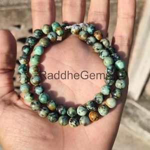 May include: A beaded necklace featuring round, turquoise-colored beads with brown and black speckles. The necklace is held in a hand and has a silver clasp. The text "RaddheGem" is visible on the necklace.