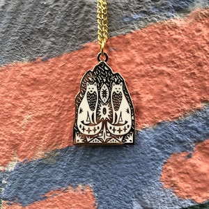 May include: A gold necklace with a pendant featuring a stylised design of three white cats sitting in a house-shaped frame. The pendant has a black and white geometric pattern.