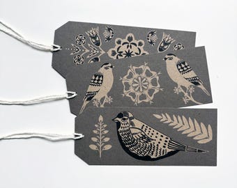 Puede incluir: Tres etiquetas de regalo grises con diseños de pájaros y flores en negro y crema. Cada etiqueta tiene una cuerda blanca.