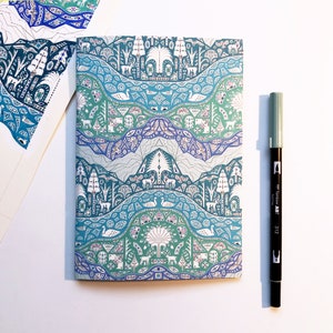 May include: A notebook with a colorful, patterned cover featuring a repeating design of trees, birds, and geometric shapes in shades of blue, green, and white. The notebook is lying on a white surface.