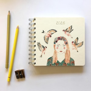 Pode incluir: Um caderno espiral com uma ilustração em aquarela de uma mulher rodeada por pássaros, o ano de 2026 está impresso acima. Dois lápis e um apontador estão ao lado do caderno.