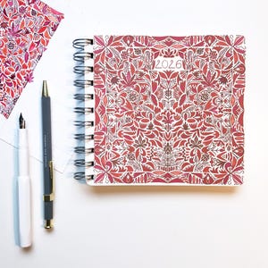 May include: A red and white spiral bound notebook with a floral pattern and the year 2026 printed on the cover. The notebook is on a white surface with two pens.