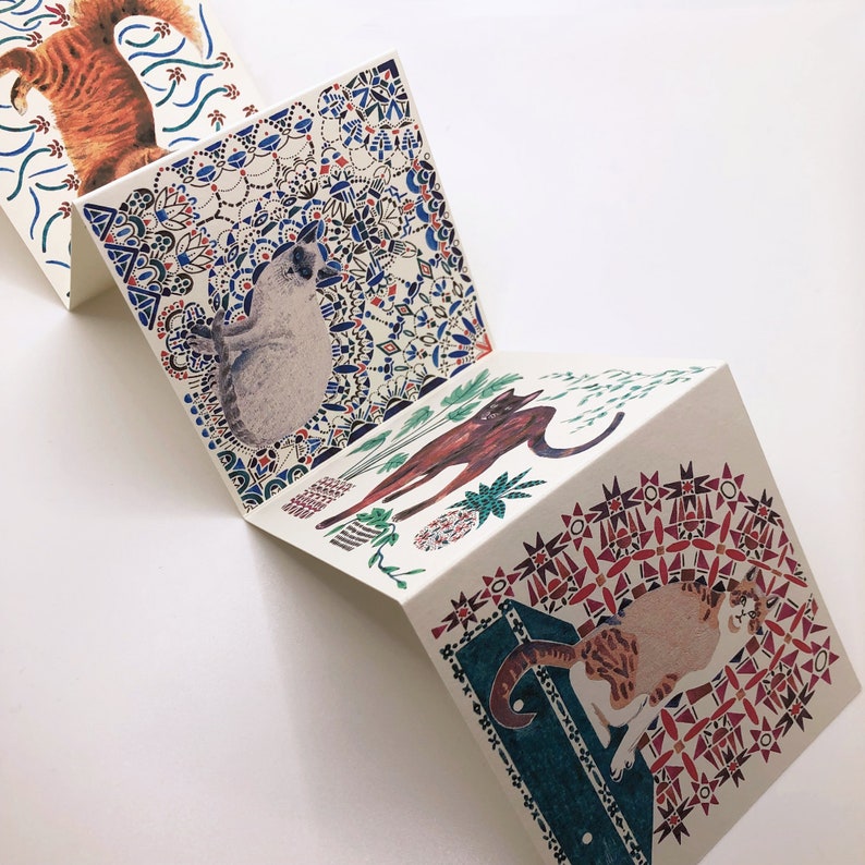 May include: A set of four folded cards with colorful patterned designs featuring cats and plants. The cards are folded in half and have a white background with blue, red, and green accents.