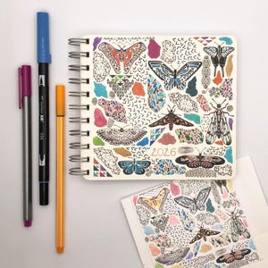 May include: A white spiral bound notebook with a colorful butterfly design. The cover features a variety of butterflies, moths, and geometric patterns. The year 2026 is printed in gold lettering on the cover.