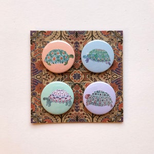 May include: Four round pinback buttons with colourful patterned turtle designs. The buttons are on a patterned background.