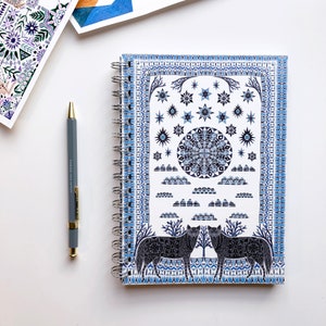 Puede incluir: Una libreta de espiral blanca con una cubierta de patrón azul y blanco que presenta un diseño de mandala, nubes, estrellas y dos lobos negros. La cubierta tiene un borde de patrones geométricos repetidos.