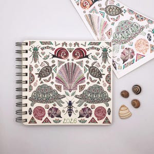 Puede incluir: Una libreta de espiral blanca con un diseño colorido que presenta tortugas, conchas marinas e insectos. La portada tiene un patrón repetitivo y el año 2026 impreso en letras doradas.