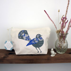Op de afbeelding: Een witte canvas toilettas met een blauw en grijs vogelontwerp. De vogel heeft een gedetailleerd patroon op zijn staartveren en een klein, rond hoofd. De tas ligt op een houten plank met een vaas met gedroogde bloemen en een klein keramiek vogeltje.