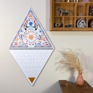 Puede incluir: Un calendario de pared en forma de diamante con un diseño geométrico colorido. La sección superior presenta un sol, pájaros y otras formas abstractas en naranja, azul y rojo. El calendario muestra el mes de agosto. La sección inferior tiene un fondo blanco con los días del mes.
