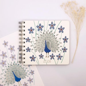 May include: A white notebook with a blue peacock design and blue flowers. The notebook has a spiral binding and the year 2026 printed on the cover.