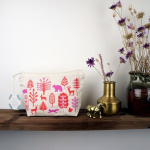 Op de afbeelding: Een witte canvas toilettas met een roze en rode print van een bos. De print bevat bomen, dieren en geometrische patronen. De tas ligt op een houten plank met een gouden vaas en een bruine keramische vaas.