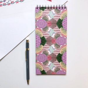 May include: A spiral bound notebook with a colourful geometric pattern in shades of pink, green, yellow, and brown. The notebook is lying on a white surface next to a grey pen.