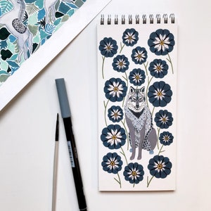 May include: A gray wolf with a geometric pattern sits in the center of a blue and white floral design. The wolf is surrounded by stylized flowers with white centers and blue petals. The background is white.