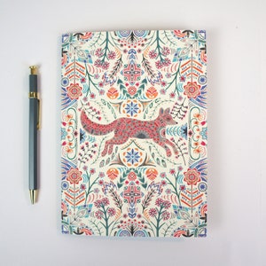Puede incluir: Un cuaderno de color crema con un patrón floral colorido y una ilustración de un zorro rojo, rosa y azul en el centro. El cuaderno tiene un borde decorativo con un patrón repetitivo de flores y hojas.