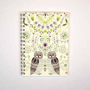 Puede incluir: Una libreta de espiral con una cubierta de color crema que presenta un diseño simétrico de búhos, flores y formas geométricas en tonos verdes, morados y marrones.