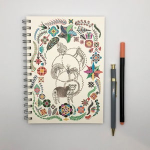 Könnte beinhalten: Ein spiralgebundenes Notizbuch mit einem farbenfrohen Design mit Tieren und Blumen. Der Einband ist weiß mit einer Vielzahl von Farben, darunter Rot, Blau, Grün, Gelb und Orange. Die Tiere sind stilisiert und umfassen ein Reh, einen Bären und einen Vogel.