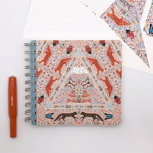 May include: A blue spiral bound planner with a colorful patterned cover featuring red foxes, butterflies, and ladybugs. The cover has a triangular design with the year 2026 printed in the center.