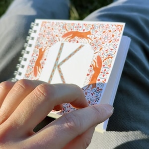 May include: A small, spiral bound notebook with a colourful cover featuring a stylised letter "K" surrounded by a border of orange foxes and floral designs.