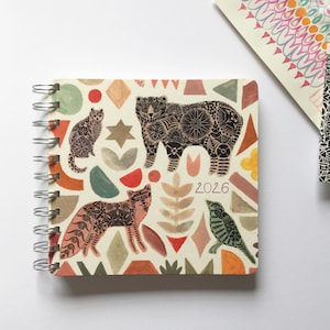 May include: A spiral bound 2026 calendar with a colourful geometric design featuring a bear, fox, cat, and bird. The cover is cream coloured with a variety of shapes and patterns in shades of red, orange, yellow, green, blue, and brown.