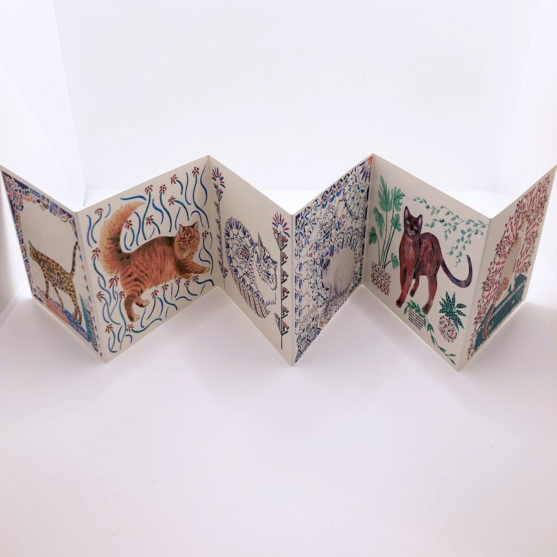 May include: A folded paper book with colorful illustrations of cats on each page. The cats are depicted in various poses and surrounded by floral and geometric patterns.