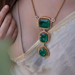 May include: A gold necklace featuring three emerald-cut green gemstones. Each gemstone is set in a gold frame and surrounded by small, clear accent stones. The necklace hangs on a delicate gold chain.
