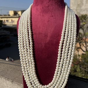 Pode incluir: Um colar de pérolas de várias voltas exibido em um busto de veludo bordô. O colar apresenta várias voltas de pérolas redondas, esbranquiçadas, caindo em cascata sobre o busto. As pérolas são de tamanho uniforme e criam um visual clássico e elegante. O fundo está desfocado.