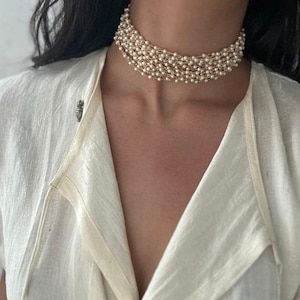 Puede incluir: Un collar gargantilla de varias hebras con pequeñas perlas blancas redondas y una delicada cadena dorada. El collar se ajusta cerca del cuello, creando un efecto de capas. La persona lleva una blusa color crema.