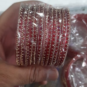 Peut inclure: Un ensemble de bracelets rigides rouge foncé avec une finition lisse et brillante, ornés de plusieurs rangées de petites pierres claires et réfléchissantes. Les bracelets sont emballés dans du plastique transparent, mettant en valeur la couleur vive.