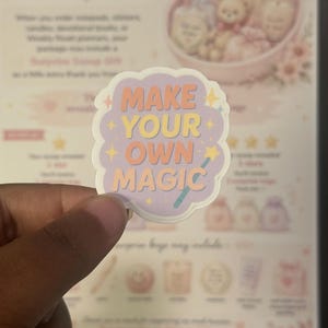 May include: A die-cut sticker with a scalloped edge featuring a light purple background and the text MAKE YOUR OWN MAGIC in orange and yellow letters. The design includes small yellow star icons and a teal wand graphic extending from the word MAGIC.