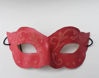May include: Red masquerade mask with glitter accents and black elastic bands. The mask features an ornate, symmetrical design with eye openings and a scalloped edge. A decorative accessory for parties and events.