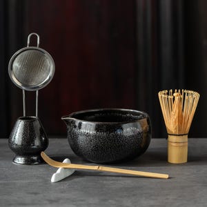 Puede incluir: Un juego de té matcha que incluye un cuenco de cerámica negra moteada con pico vertedor, un batidor de bambú, una cuchara de bambú sobre un soporte de cerámica blanca y un colador de metal sobre una base de cerámica negra moteada.