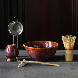 May include: A matcha tea set featuring a glazed ceramic bowl with a pouring spout, a bamboo whisk, a bamboo scoop, a ceramic tea caddy, and a stainless steel tea strainer. The bowl and caddy have a mottled red and purple glaze.
