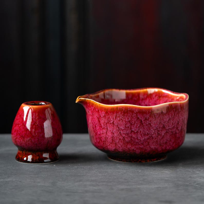 May include: A ceramic matcha whisk holder and a matching pouring bowl, both featuring a vibrant magenta and deep red crystalline glaze with a glossy finish. The bowl has a wide, scalloped rim and a small spout for pouring liquids with precision.
