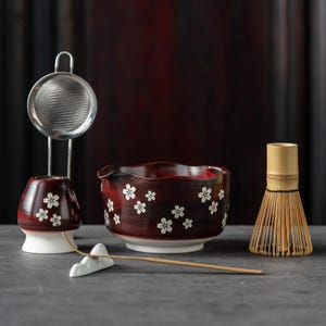Op de afbeelding: Een Japanse matcha-theeset met een donkerrode keramische kom en beker met witte bloemmotieven. Inclusief een bamboe klopper, een theelepel, een keramische rust en een roestvrijstalen thee-ei.