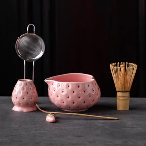 Könnte beinhalten: Eine rosa Keramik-Matchaschale und ein Besenhalter in Erdbeerform mit roten, punktförmigen Vertiefungen, ein Bambus-Matchabesen, ein Metallsieb und ein langer Bambuslöffel auf einer kleinen, erdbeerförmigen Keramikablage.