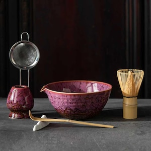 May include: A matcha tea set featuring a large, deep purple bowl with a spout, a bamboo whisk, a scoop, a small ceramic holder, and a stainless steel strainer. The set is designed for preparing matcha tea.