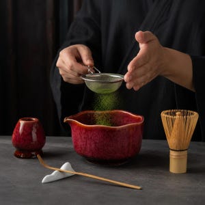 May include: A matcha tea set featuring a deep red, textured ceramic bowl with a pouring spout, a matching red whisk holder, a bamboo whisk, and a bamboo tea scoop resting on a white ceramic stand. Green matcha powder is being sifted into the bowl.