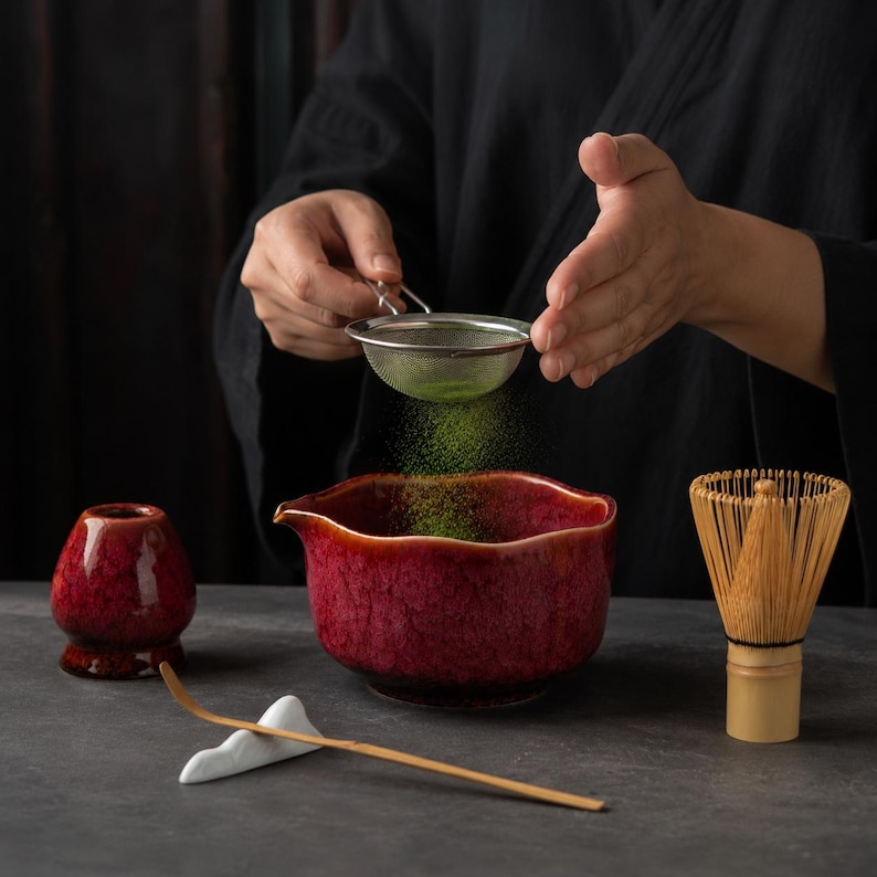 May include: A matcha tea set featuring a deep red, textured ceramic bowl with a pouring spout, a matching red whisk holder, a bamboo whisk, and a bamboo tea scoop resting on a white ceramic stand. Green matcha powder is being sifted into the bowl.
